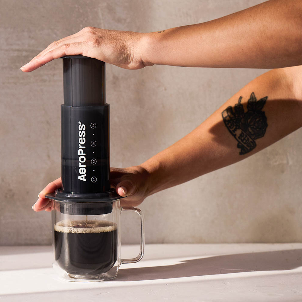 Person using an Aeropress coffee maker on a kitchen counter with a plant in the background.