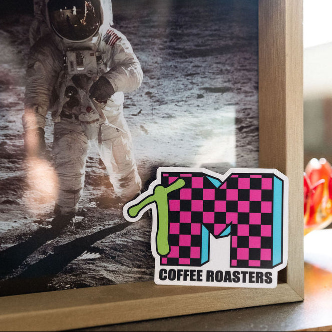 Checkered logo with 'TM Coffee Roasters' text on a surface with an astronaut image in the background