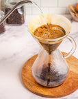 Coffee being poured into a glass carafe on a marble countertop with coffee beans and a plant in the background.