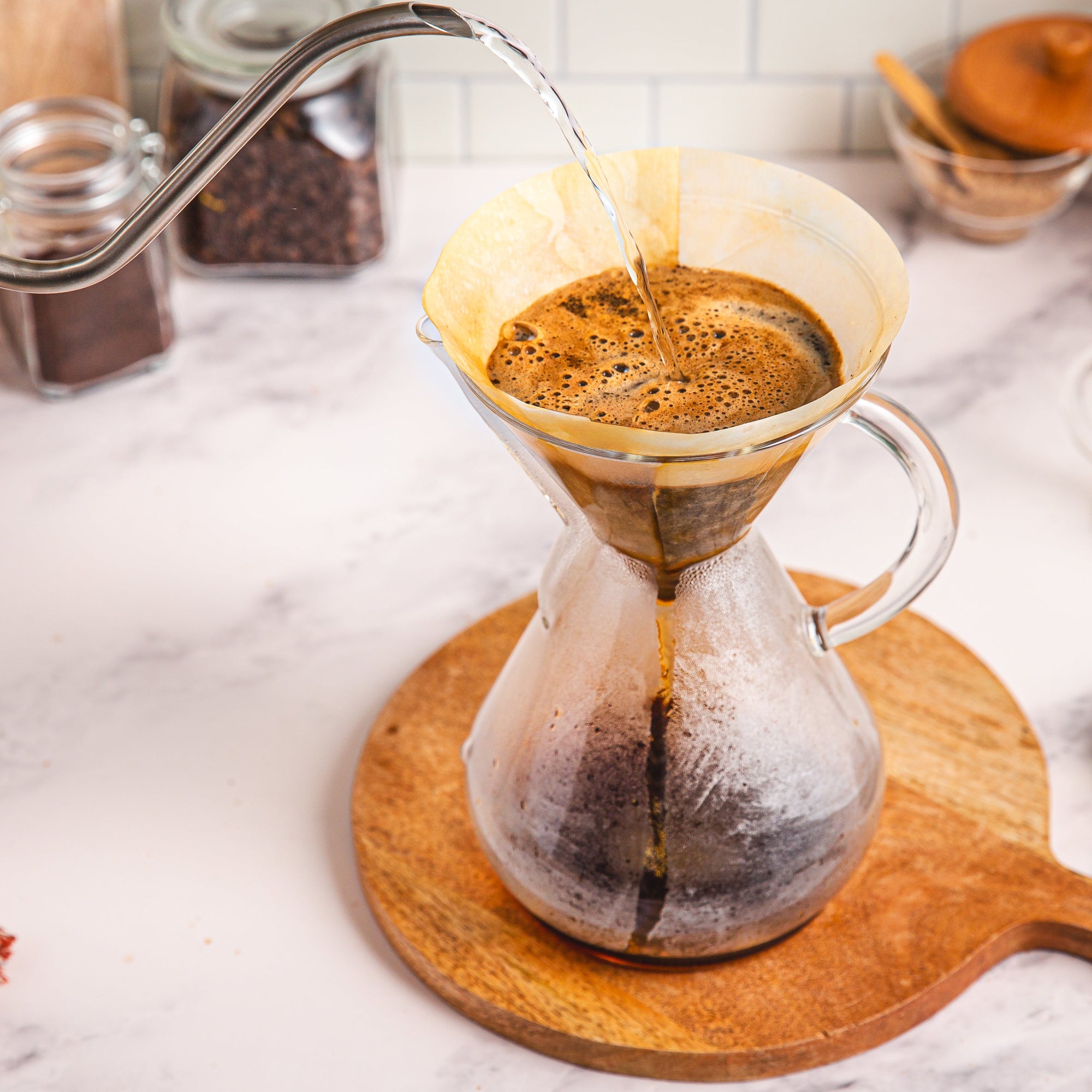 Coffee being poured into a glass carafe on a marble countertop with coffee beans and a plant in the background.